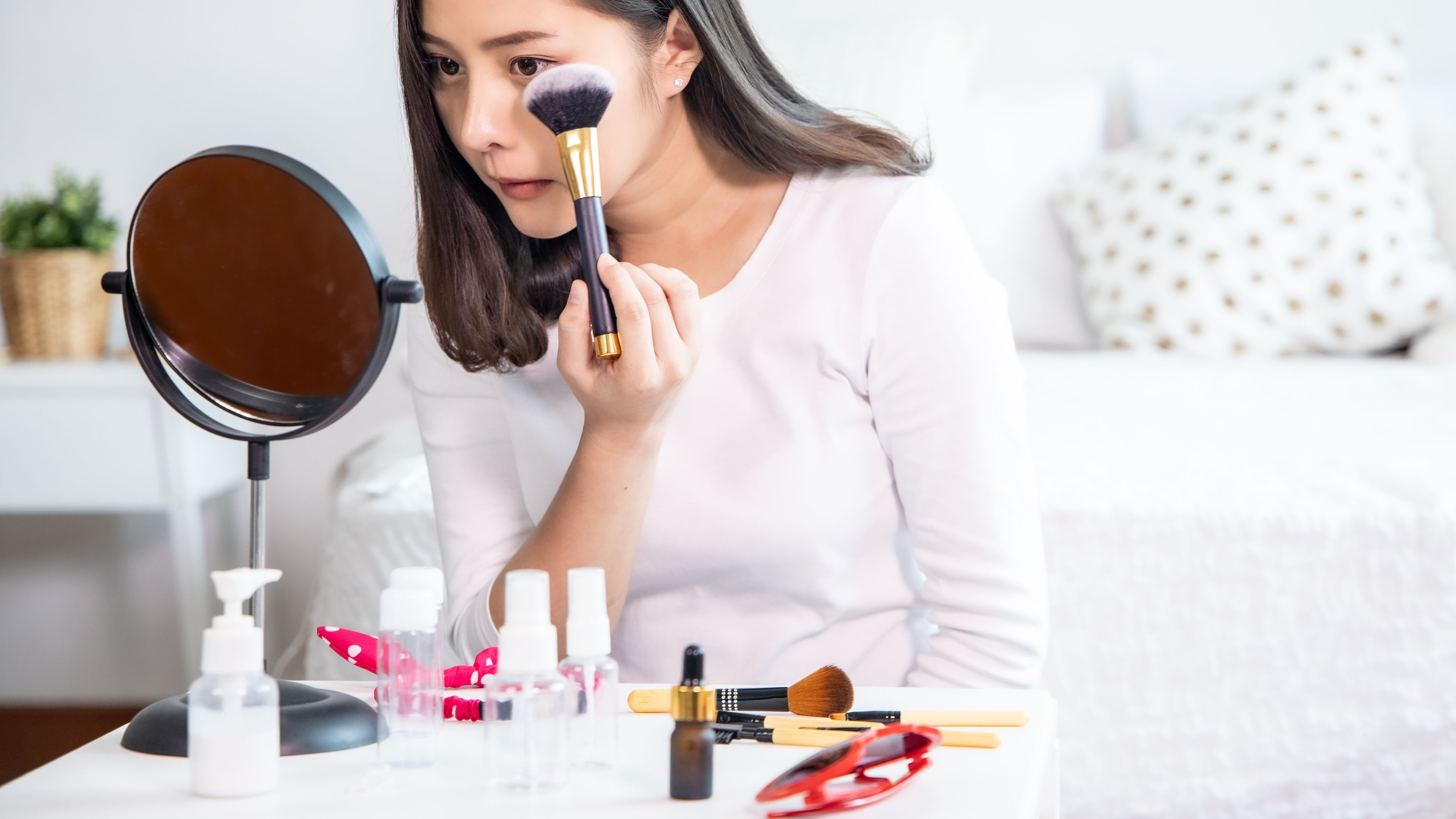 A young woman applying makeup with a brush while looking into a round mirror on a white table with cosmetic products.